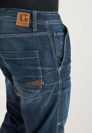 Mørkeblå denimjeans med et læderpatch-label, kontrasterende syninger, for- og baglommer samt lynlås. Synlige slid detaljer.