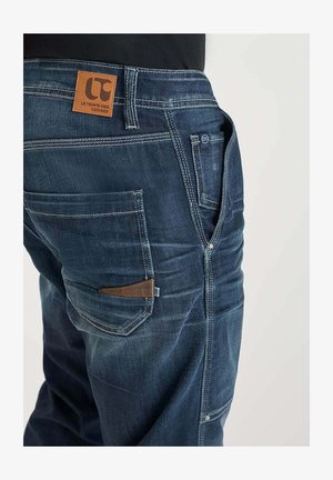 Donkerblauwe denim jeans met een leren patchlabel, contrasterende stiksels, voor- en achterzakken, en een ritssluiting. Zichtbare slijtage details.