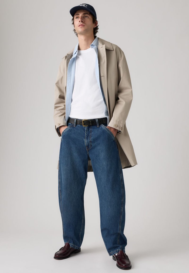 Gabardina beige sobre una camisa azul claro y una camiseta blanca. Vaqueros azules holgados y zapatos de cuero oscuro. La gorra añade un toque casual.