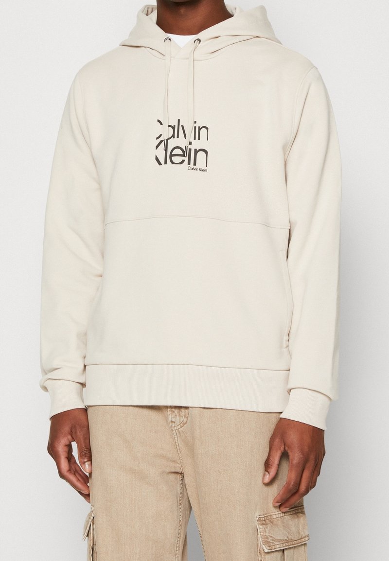 Hoodie beige en tissu doux, avec une poche kangourou à l'avant et un logo noir contrastant au centre. Poche kangourou à l'avant.
