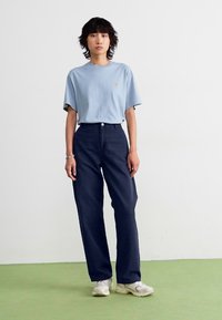 T-shirt bleu clair à manches courtes avec un petit logo, associé à un pantalon large bleu marine et des baskets blanches sur un sol vert.