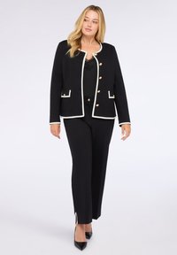 Sort skræddersyet jakke med hvid trim og guldknapper, der har to forlommer. Båret over en sort top og parret med sorte bukser.