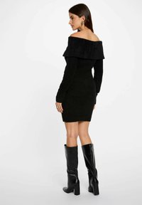 Vestido de punto negro sin hombros con mangas largas y silueta ajustada, combinado con botas altas de tacón negro brillante.