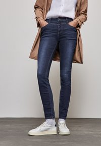 Mörkblå denim skinny jeans med framfickor, matchade med vita sneakers och ljusgrå strumpor, kompletterat med en beige trenchcoat.