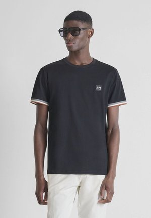 Muškarac obučen u crnu majicu kratkih rukava s prugastim rubovima rukava i malim logotipom, bijele hlače i velike tamne sunčane naočale, na jednobojnoj pozadini.