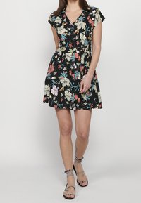 Vestido envolvente floral con fondo negro, mangas cortas, escote en V y detalle de cintura anudada. Los colores incluyen flores en tonos rosa, azul y blanco.