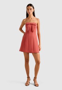 Frau in einem schulterfreien korallenfarbenen Minikleid mit großem Schleifendetail, die mit einer Hand auf der Hüfte steht und schwarze High-Heels trägt.