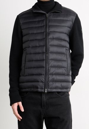 Mies, joka käyttää mustaa tikattua vetoketjullista liiviä mustan neulesweaterin ja mustien housujen päällä, kuvattuna olkapäistä lanteisiin yksiväristä taustaa vasten.