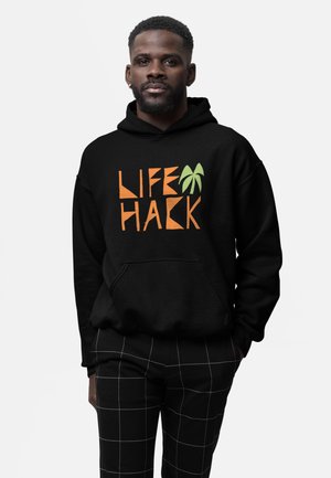 Mann trägt schwarzen Hoodie mit dem Schriftzug "LIFE HACK" und grünem Palmen-Motiv, kombiniert mit schwarzen Hosen mit weißen Gitterlinien.