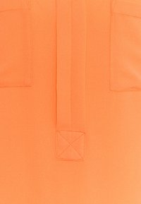 Blouse orange avec une texture lisse, un détail de col et deux poches poitrine ornées de surpiqûres carrées. Tissu léger.