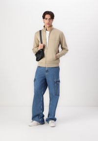Beige Zip-Jacke mit hohem Kragen, über einem weißen T-Shirt getragen. Lockere blaue Cargo-Jeans mit Taschen und weißen Turnschuhen. Schwarze Umhängetasche.