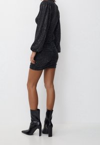 Kvinna i en svart paljettklänning med lång ärm och mini längd, kombinerad med svarta glänsande klackboots som går upp till ankeln, stående mot en enkel vit bakgrund.