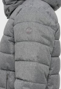 Grå puffjacka med ett texturerat tyg, kviltad design och en cirkulär logotyp-patch på överarmen. Har fickor med dragkedja.