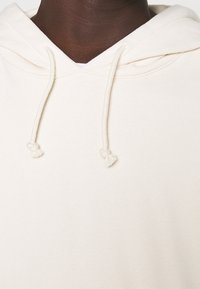 Krämfärgad hoodie gjord av mjukt tyg med en avslappnad passform. Har en huva med dragsko och knutade ändar samt en strukturerad yta.