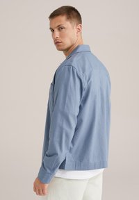 Camisa de chambray azul con un corte relajado, mangas largas, un collar y un bolsillo en el pecho, llevada sobre una camiseta blanca y pantalones claros.