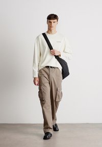 Helles cremefarbenes Langarmhemd mit dezentem Logo, kombiniert mit beigen Cargo-Hosen mit mehreren Taschen. Schwarze Umhängetasche und Schuhe.