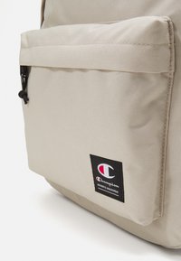 Beige canvas backpack with a front pocket, adjustable drawstring, and a black Champion logo patch. Textured fabric with stitched seams.