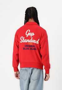 Crvena dukserica s natpisom "Gap Standard 1969 ORIGINAL BLUE JEANS" u bijelom i plavom tekstu na leđima. Ima opušteni kroj i rebraste manžete.