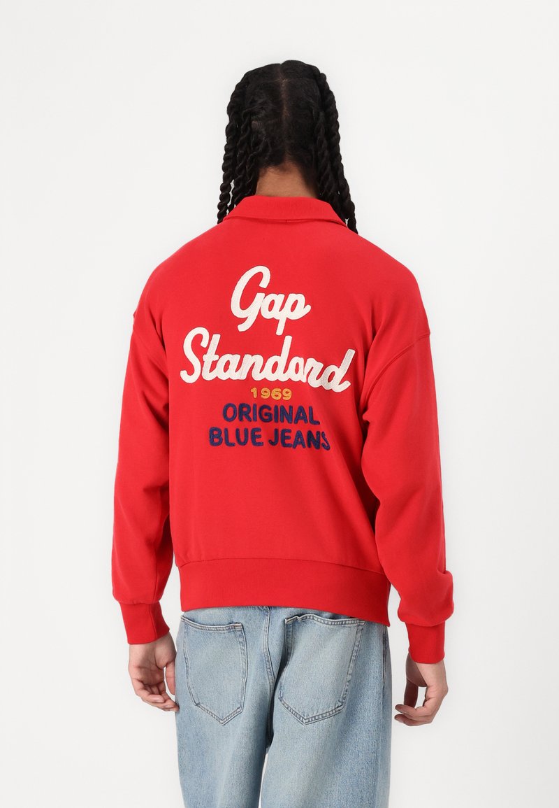 Crvena dukserica s natpisom "Gap Standard 1969 ORIGINAL BLUE JEANS" u bijelom i plavom tekstu na leđima. Ima opušteni kroj i rebraste manžete.