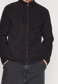 Chaqueta de forro polar negra con cuello alto, cierre frontal con cremallera y bolsillos laterales. Textura suave y diseño minimalista.