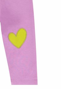 Leggings rosas con un parche de corazón amarillo en la pierna izquierda. Están hechos de una tela suave con una textura lisa, terminando en el tobillo.
