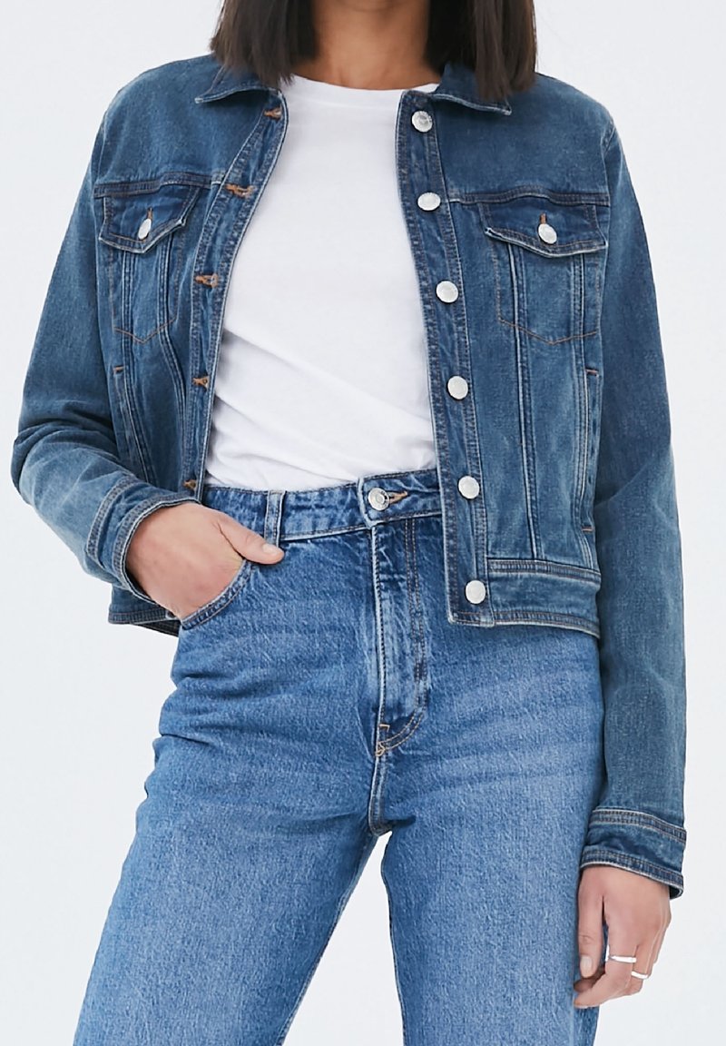 Femme portant une veste en denim bleue sur un t-shirt blanc et un jean en denim bleu taille haute, avec une main dans la poche et des bagues aux doigts.