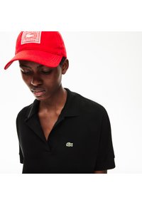 Svart polo t-shirt med en grön krokodil-logotyp på vänster bröst, ihopparad med en ljus röd basebollkeps. Tyget verkar slätt och strukturerat.