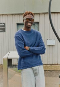Jeune homme portant des lunettes de soleil teintées orange et un pull bleu, debout les bras croisés et souriant à l'extérieur près d'une poubelle en métal et d'un mur de bâtiment.