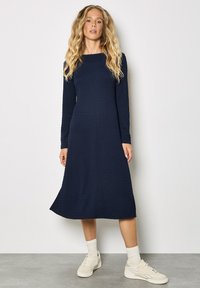 Vestido azul marino de manga larga con tela texturizada, silueta en línea A, escote redondo y longitud hasta la mitad de la pantorrilla, combinado con zapatillas blancas.