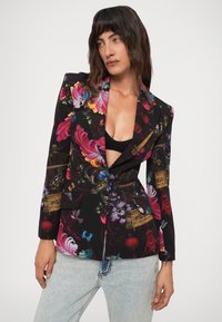 Persona con capelli scuri di media lunghezza che indossa un blazer nero con un motivo floreale e di uccelli dai colori vivaci, sopra una maglietta nera e jeans azzurri chiari, in posa.