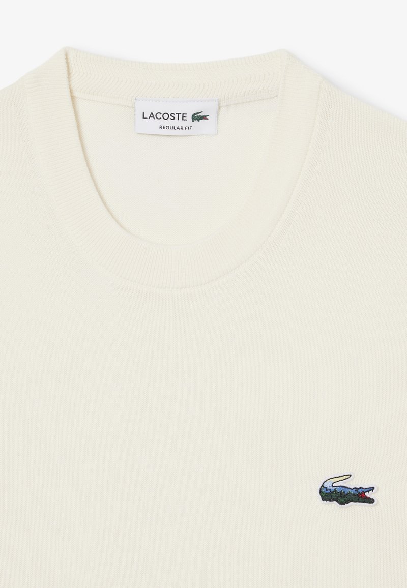 T-shirt en coton couleur crème avec un col rond, comportant un petit logo de crocodile brodé sur le côté gauche. Design à coupe régulière.