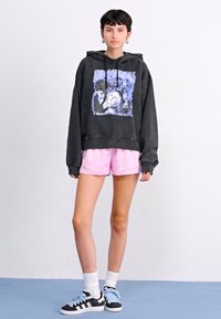 Felpa con cappuccio grigio scuro con stampa grafica, abbinata a pantaloni corti rosa chiaro. Indossata con sneakers nere e blu e calzini bianchi alla caviglia.