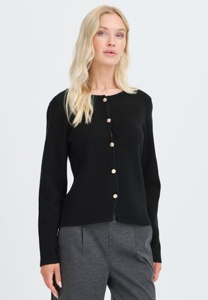 Femme aux longs cheveux blonds porte un cardigan noir boutonné et un pantalon gris taille haute sur un fond clair uni.
