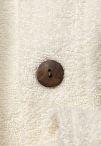 Tessuto color crema con una superficie texture e soffice, e un bottone in legno scuro con un foro centrale. Aspetto morbido e accogliente.