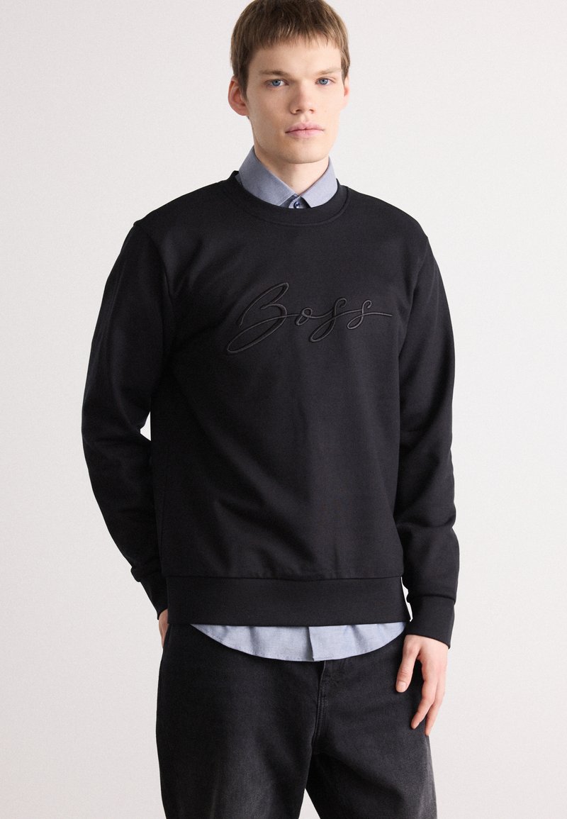 Sweatshirt preto com decote redondo e logótipo "Boss" em relevo na parte da frente. Tecido em mistura de algodão, mangas longas e punhos canelados. Sobreposto a uma camisa azul clara.