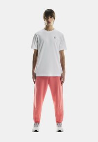 Vit t-shirt med kort ärm och svart logotyp, kombinerad med korallfärgade joggers med elastiska muddar. Modellen bär grå löparskor.