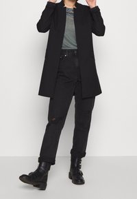 Abrigo largo negro de corte recto, combinado con vaqueros oscuros con un detalle roto y botines negros con hebillas. Camiseta gris debajo.