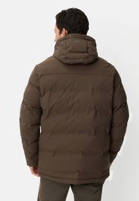 Brun pufferjakke med hætte, der har en glat tekstur, quiltet design og synlige stikninger. Bagsidevisning, logo på hætten.