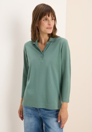 Polo en vert clair à manches longues avec une patte de boutonnage à deux boutons, texture douce et coupe décontractée, associé à un jean bleu clair.
