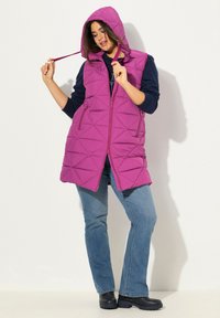 Fuchsia pufferväst med huva, med en geometrisk kviltad design, sidofickor och dragkedjestängning. Bärs över en marinblå topp och jeans.