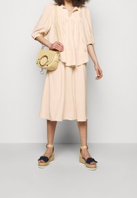 Blouse rose pâle avec détails froncés, accompagnée d'une jupe midi fluide. Sandales compensées avec franges bleu marine. Sac crossbody jaune.