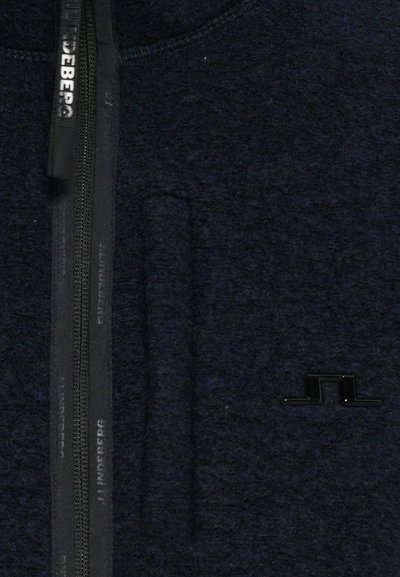 Marineblaues Fleece-Jackett mit einem Reißverschluss an der Vorderseite, mit einem schwarzen Logo und strukturiertem Material. Enthält eine Vordertasche mit Nähdetails.