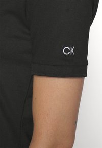 Gros plan sur le haut du bras d’une personne portant un t-shirt noir à manches courtes avec des lettres "CK" blanches brodées sur l’ourlet de la manche.