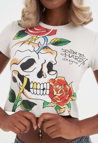 Afskåret creme T-shirt med grafik af et kranium med roser, grønne blade og farverige aksenter; blødt bomuldsstof.