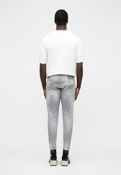 Homme debout de dos portant une chemise blanche à manches courtes, un jean gris clair slim et des bottines à talons noires.