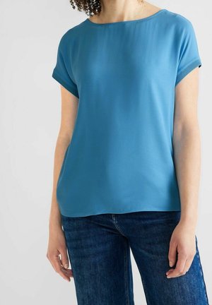 Femme portant un haut bleu uni à manches courtes et un jean bleu foncé sur un fond gris clair.