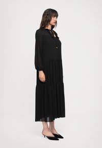 Abito nero a maniche lunghe con un design a strati, scollatura arricciata, dettaglio annodato al colletto e un materiale leggero e fluido.