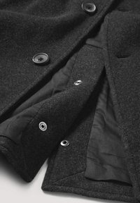 Schwarzer Wollmantel mit weicher Textur, Knopfverschluss und einem kontrastierenden schwarzen Futter mit sichtbaren Druckknöpfen.