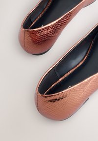 Ballerines en or rose métallisé avec une texture peau de serpent et un intérieur noir lisse, dotées d'un bout arrondi et d'un design minimaliste.