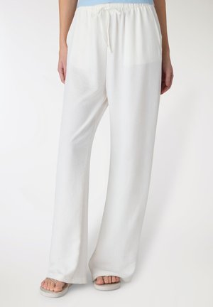 Persona che indossa pantaloni bianchi larghi con coulisse e sandali, in piedi davanti a uno sfondo chiaro e semplice.